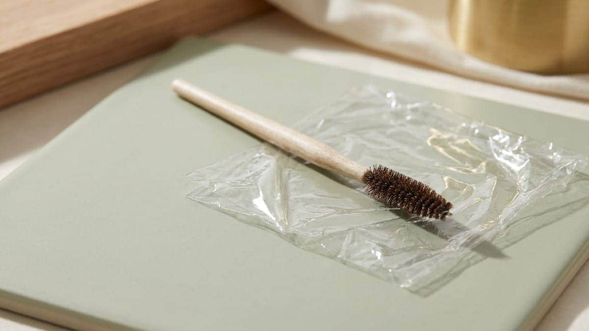 Brosse à sourcils à manche en bois clair posée sur un carré de film étirable froissé, sur un carreau de céramique vert sauge