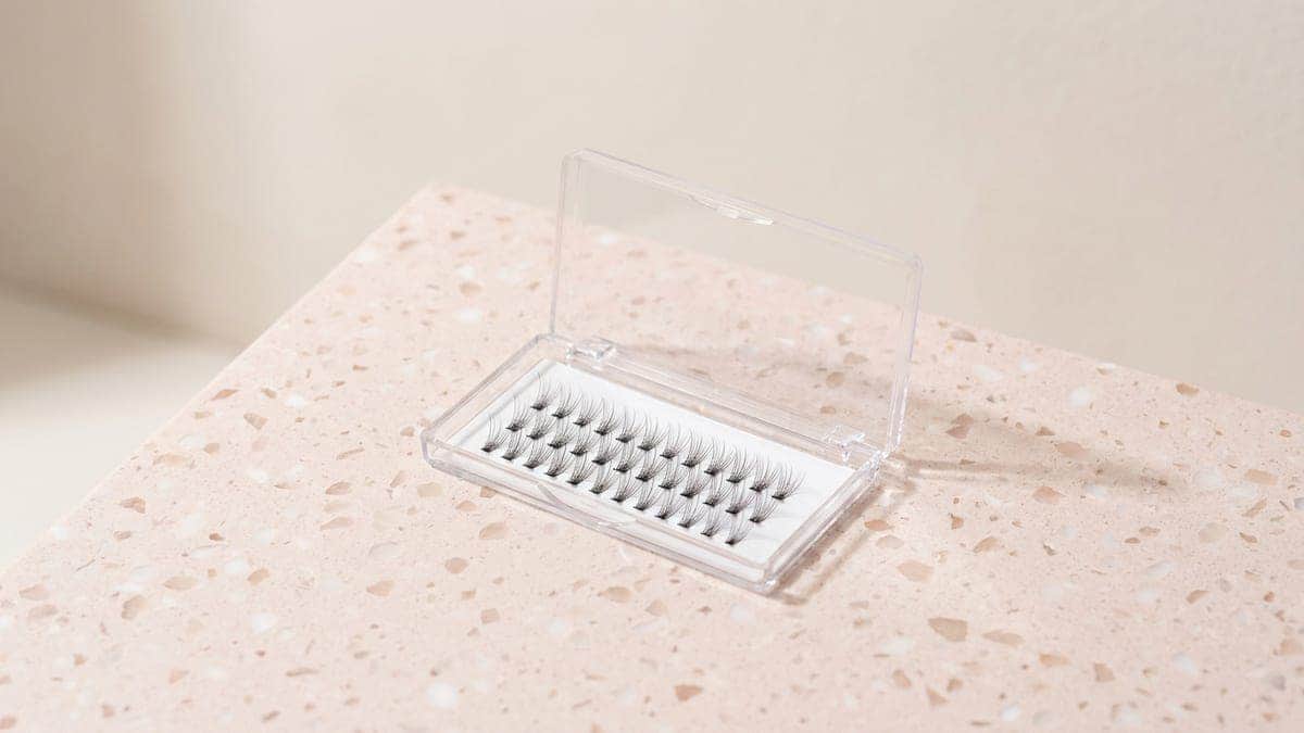 Boîtier transparent ouvert contenant des rangées de petites grappes de cils noirs, posé sur un plan en terrazzo rose