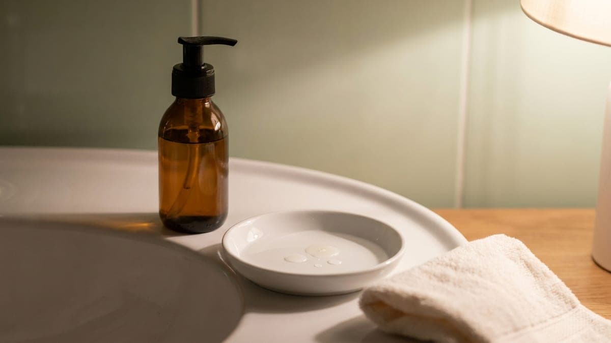 Flacon pompe d'huile démaquillante en verre ambré posé au bord d'une vasque en céramique blanche, à côté d'une petite coupelle où l'huile mêlée d'eau devient laiteuse