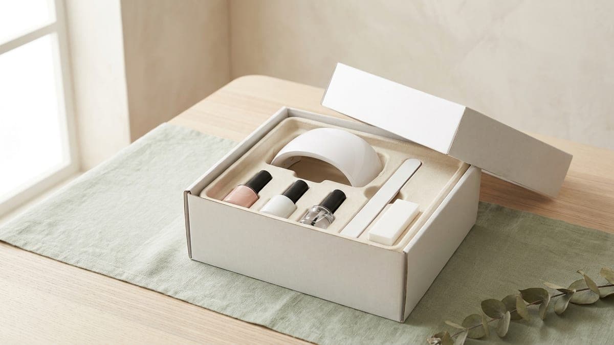 Coffret blanc ouvert posé sur un linge vert sauge : une petite lampe LED pour ongles, trois flacons de vernis semi-permanent nude, blanc et transparent, une lime et un bloc polissoir