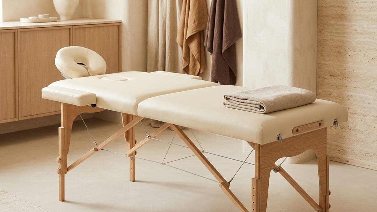 Table de massage pliante en bois clair à plateau crème, têtière et câbles de maintien visibles, dépliée dans une cabine lumineuse