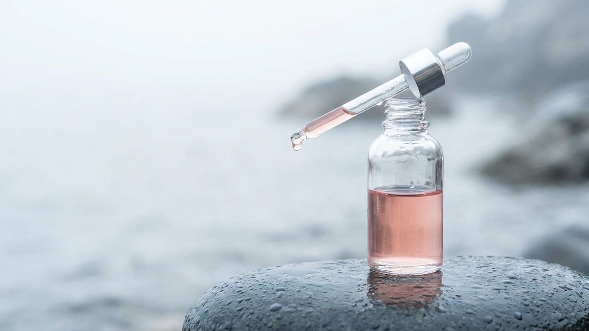 Flacon compte-gouttes de sérum rose pâle posé sur une pierre grise mouillée, une goutte suspendue au bout de la pipette