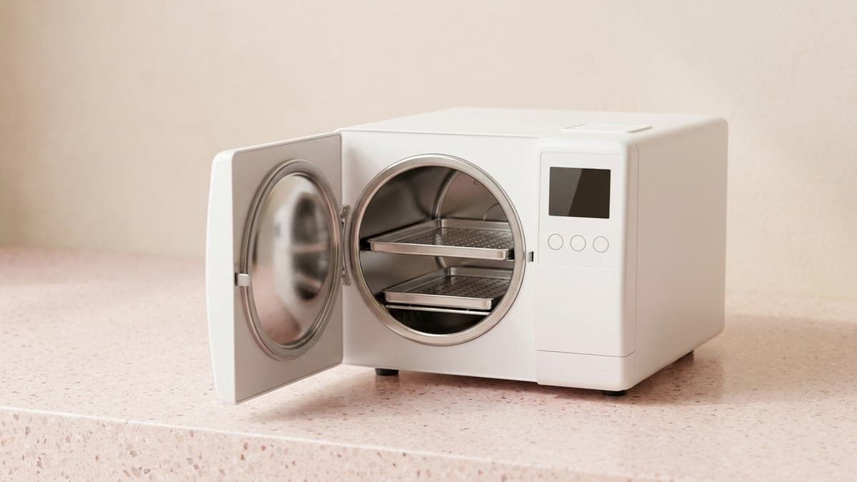 Autoclave de table blanc, porte ouverte sur une cuve ronde en inox et deux plateaux perforés
