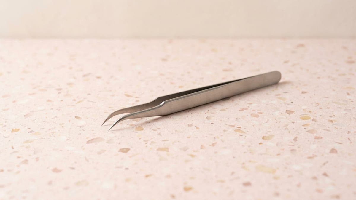 Pince d'extension de cils en acier inoxydable aux pointes fines légèrement courbées, posée en diagonale sur un plan en terrazzo rose