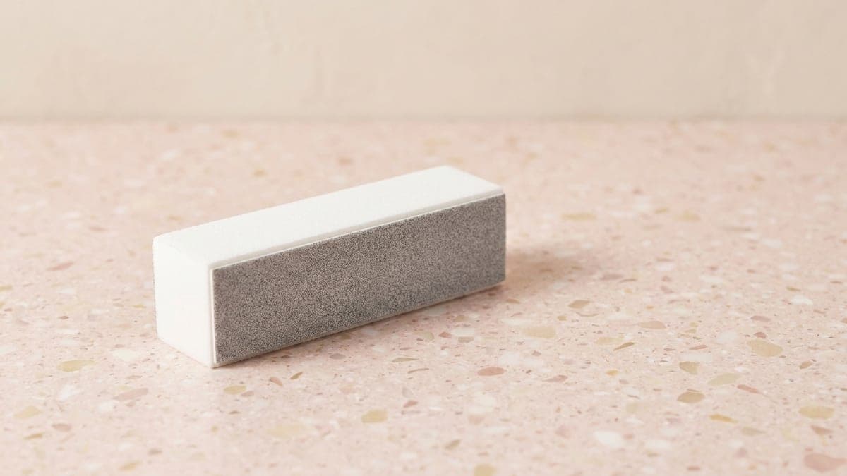 Bloc polissoir blanc pour ongles posé en biais sur un plan de travail rose poudré
