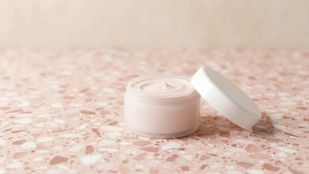Petit pot blanc ouvert de crème rose pâle, son couvercle posé à côté