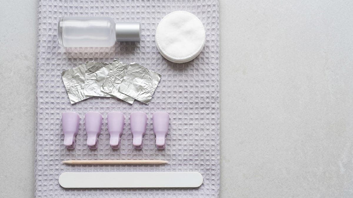 Matériel de dépose du semi-permanent posé sur une serviette gris clair : flacon de dissolvant, disques de coton, carrés de papier aluminium, cinq pinces de dépose lilas, un bâtonnet de buis et une lime blanche