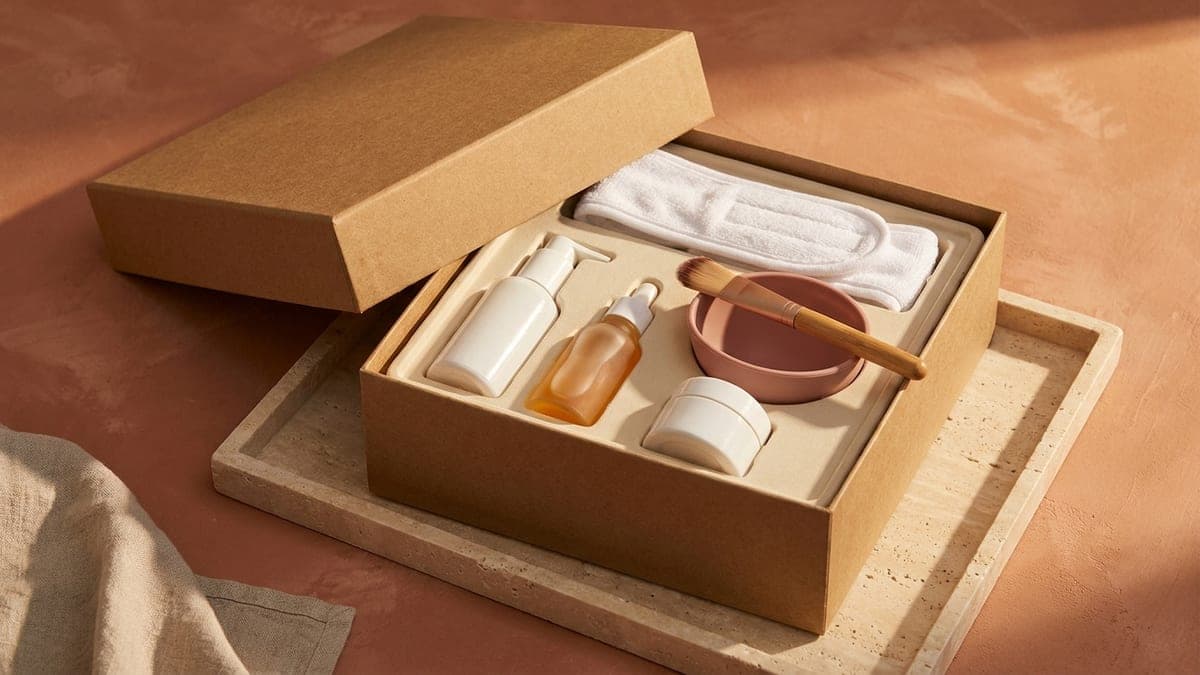 Coffret en carton kraft ouvert sur un plateau en travertin : flacon pompe de nettoyant, flacon de sérum à pipette, pot de crème, bol à masque rose poudré avec son pinceau et bandeau éponge blanc