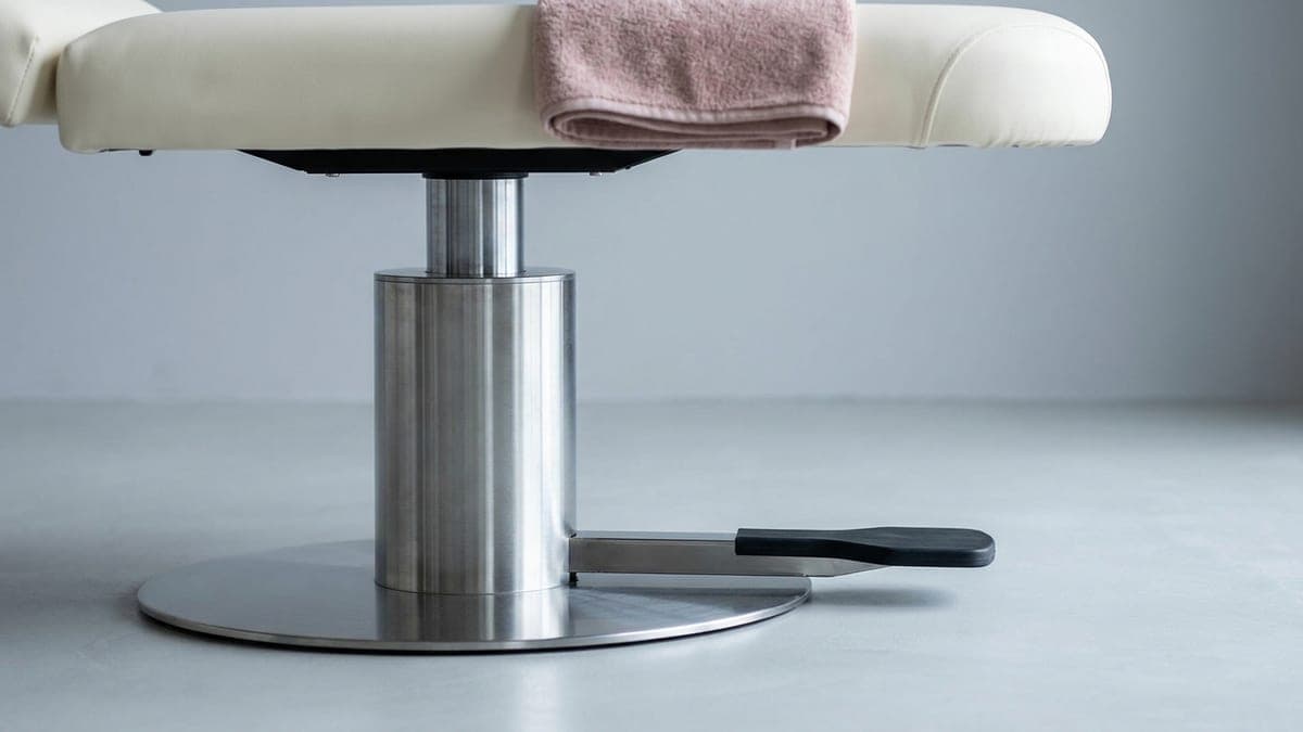 Pied d'une table de soin vu au ras du sol : colonne de levage chromée sur socle rond, large pédale noire, plateau crème et serviette rose pâle