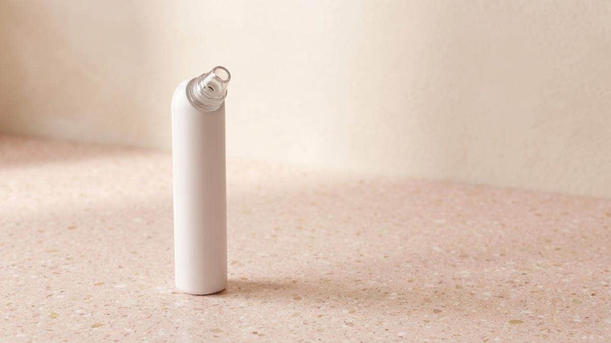 Aspirateur de points noirs blanc et fin, embout d'aspiration transparent au sommet, posé debout sur un plan en terrazzo rose
