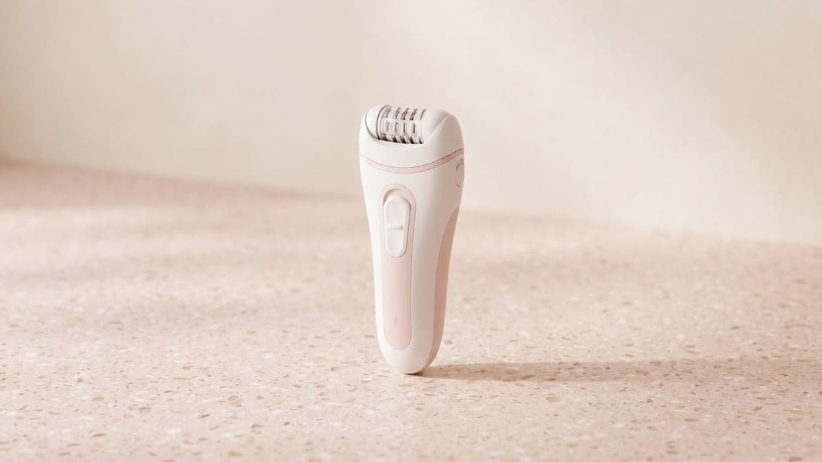 Épilateur électrique blanc et rose pâle, debout sur un plan en terrazzo rose, tête à pinces rotatives visible